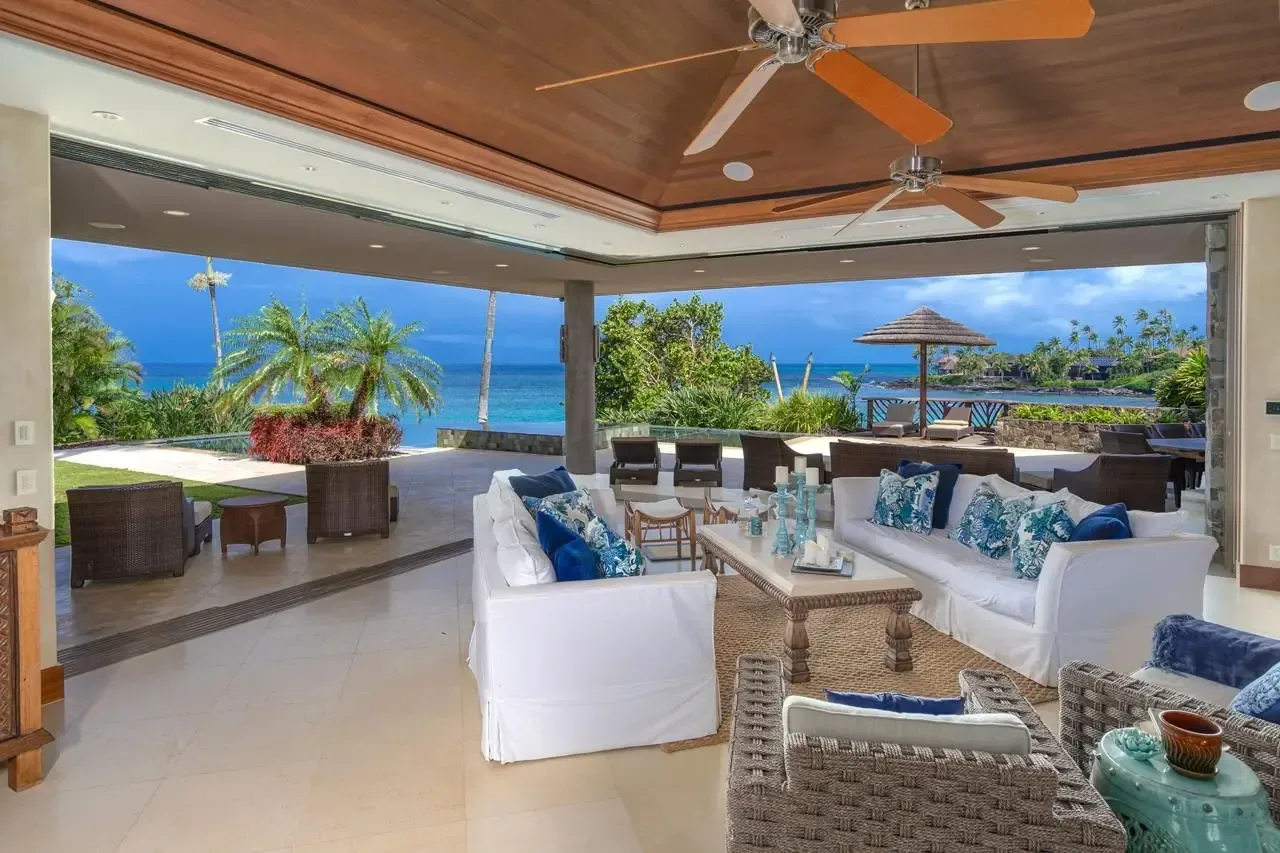 Open-air oceanfront island estate living room with a teak ceiling, white sofas, and an infinity pool overlooking the coastline.