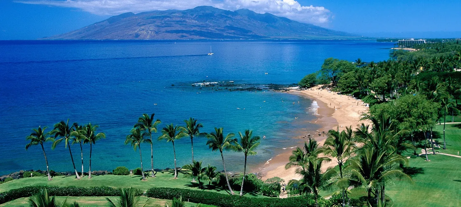 Golden crescent beach and turquoise bay on Maui's south shore, the West Maui Mountains rising across the water, palms and lawn in the foreground.
